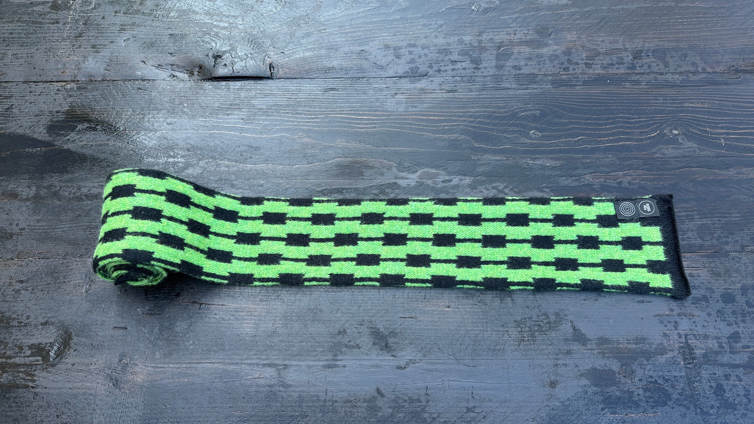 Green and black Pulse-pattern merino lambswool skinny scarf, partially unrolled on dark wood