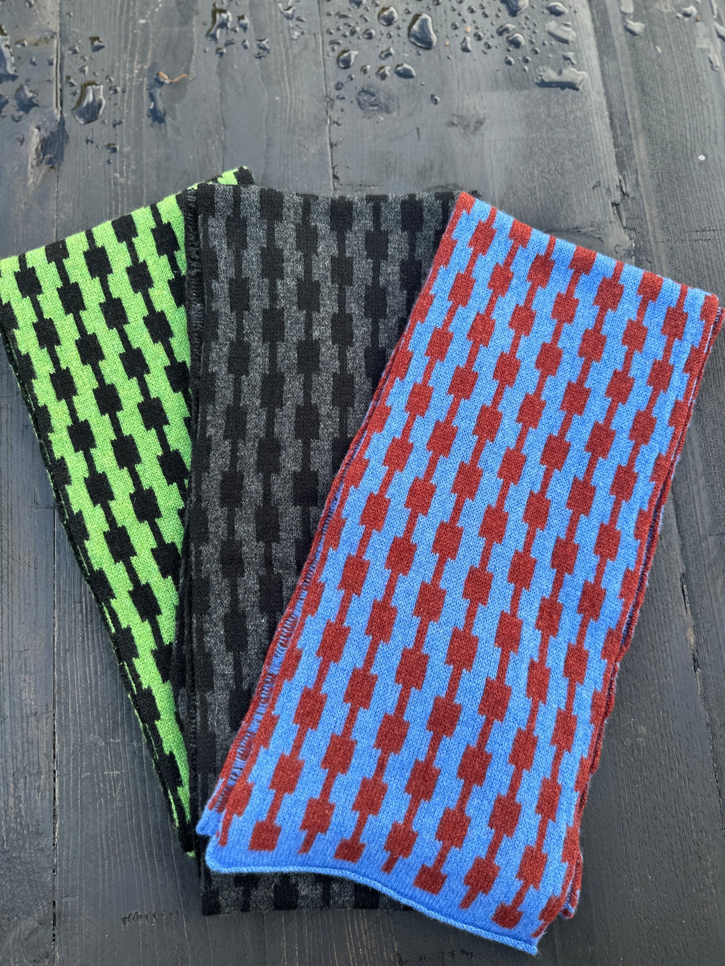 Three different coloured hand crafted merino lambswool geometric patterned Pulse scarves fanned out on a grey wooden table.