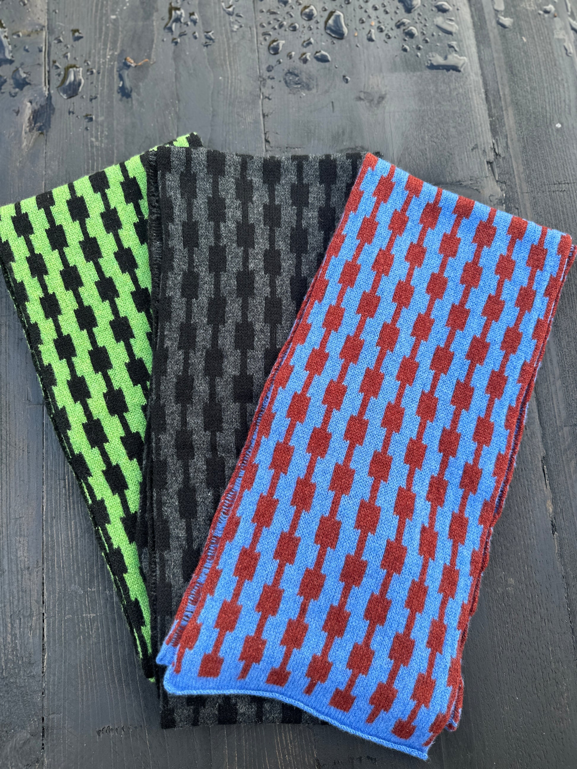 Three different coloured hand crafted merino lambswool geometric patterned Pulse scarves fanned out on a grey wooden table.