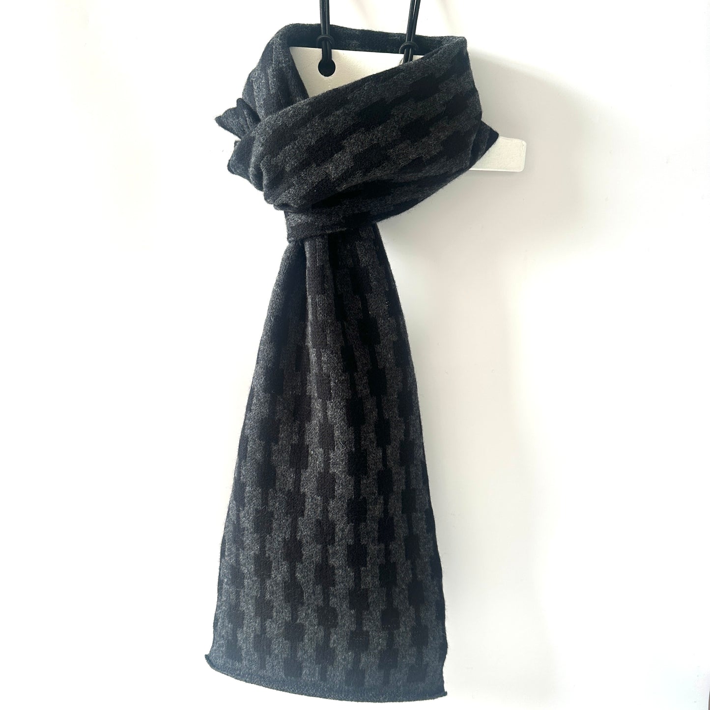 Black and charcoal grey handcrafted Merino Lambswool geometric patterned Pulse scarf on a white hanger against a white background.  Pom Shanty Knitwear. 