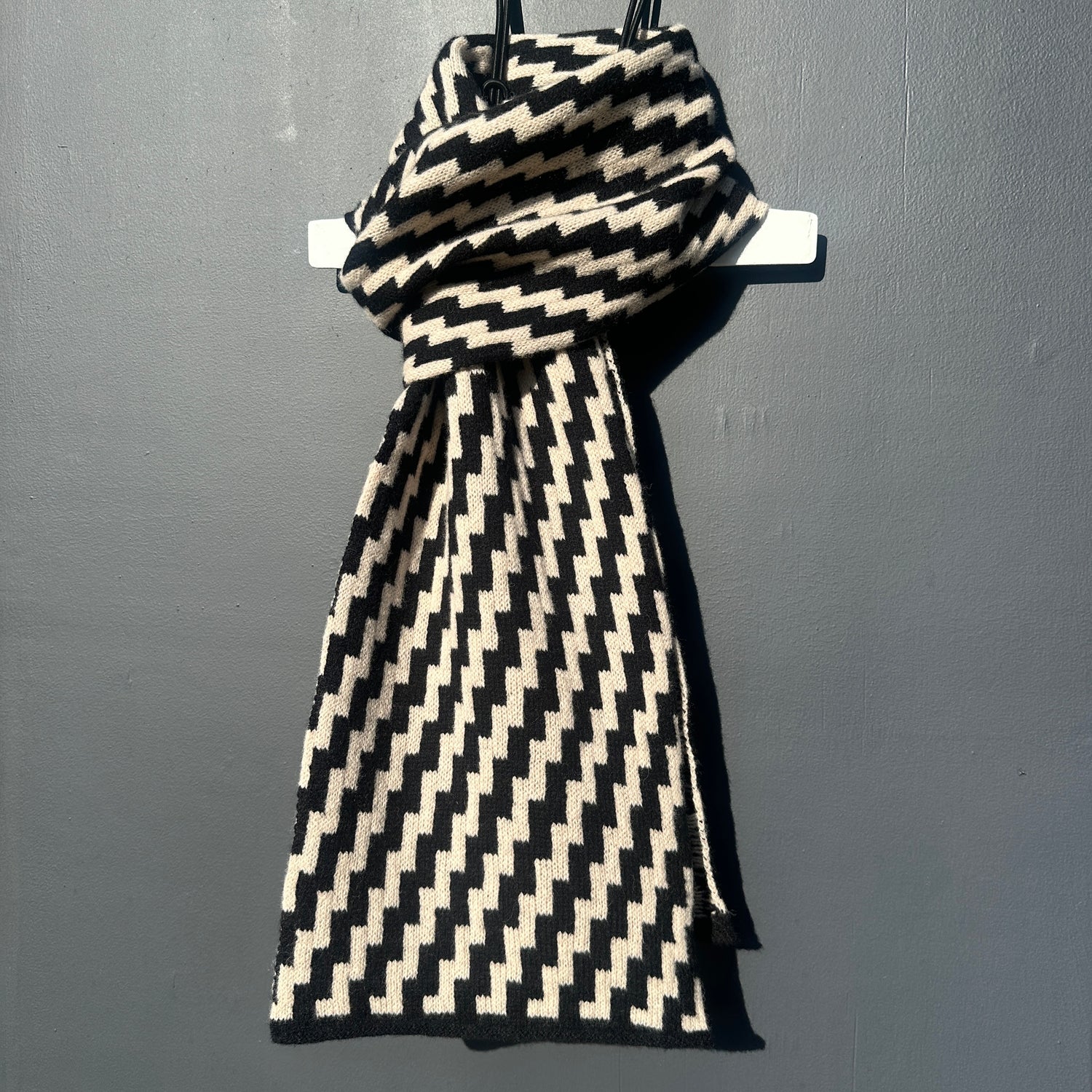 rick rack geometric patterned black and oatmeal merino lambswool scarf tied on a hanger on a rod against a grey background. Pom Shanty Knitwear.