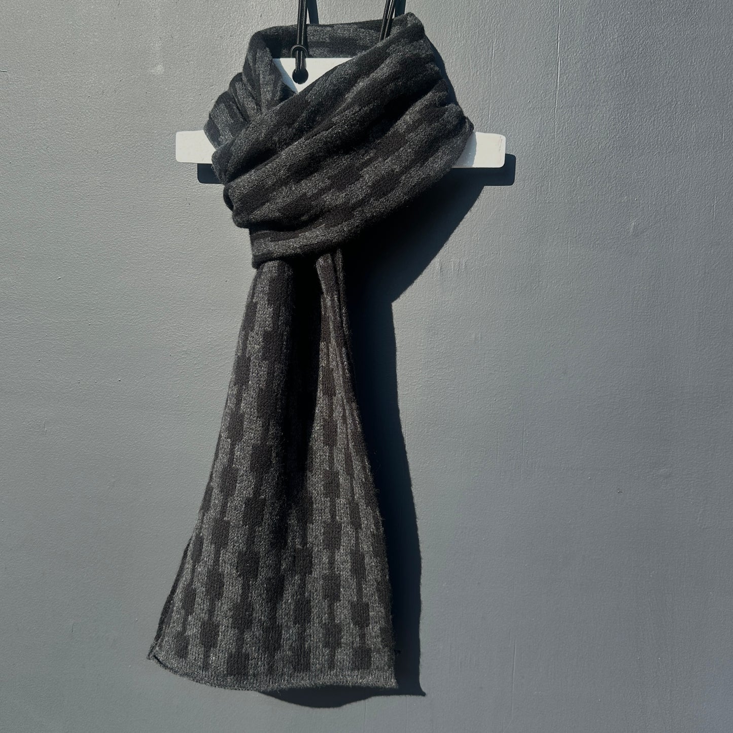 Black and charcoal grey handcrafted Merino Lambswool geometric patterned Pulse scarf on a white hanger against a grey background.  Pom Shanty Knitwear. 