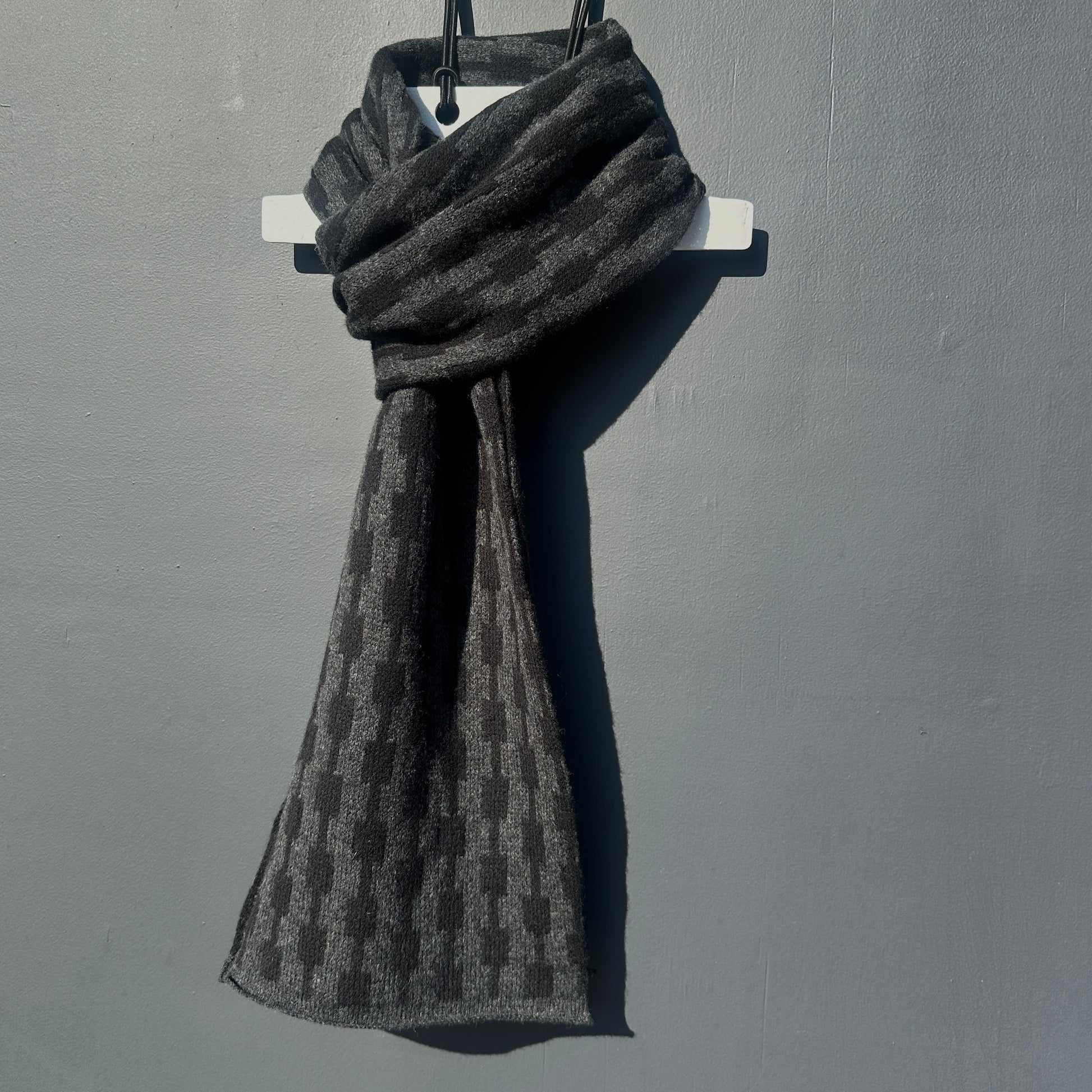 Black and charcoal grey handcrafted Merino Lambswool geometric patterned Pulse scarf on a white hanger against a grey background.  Pom Shanty Knitwear. 