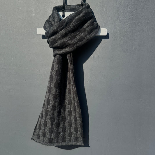 Black and charcoal grey handcrafted Merino Lambswool geometric patterned Pulse scarf on a white hanger against a grey background.  Pom Shanty Knitwear. 
