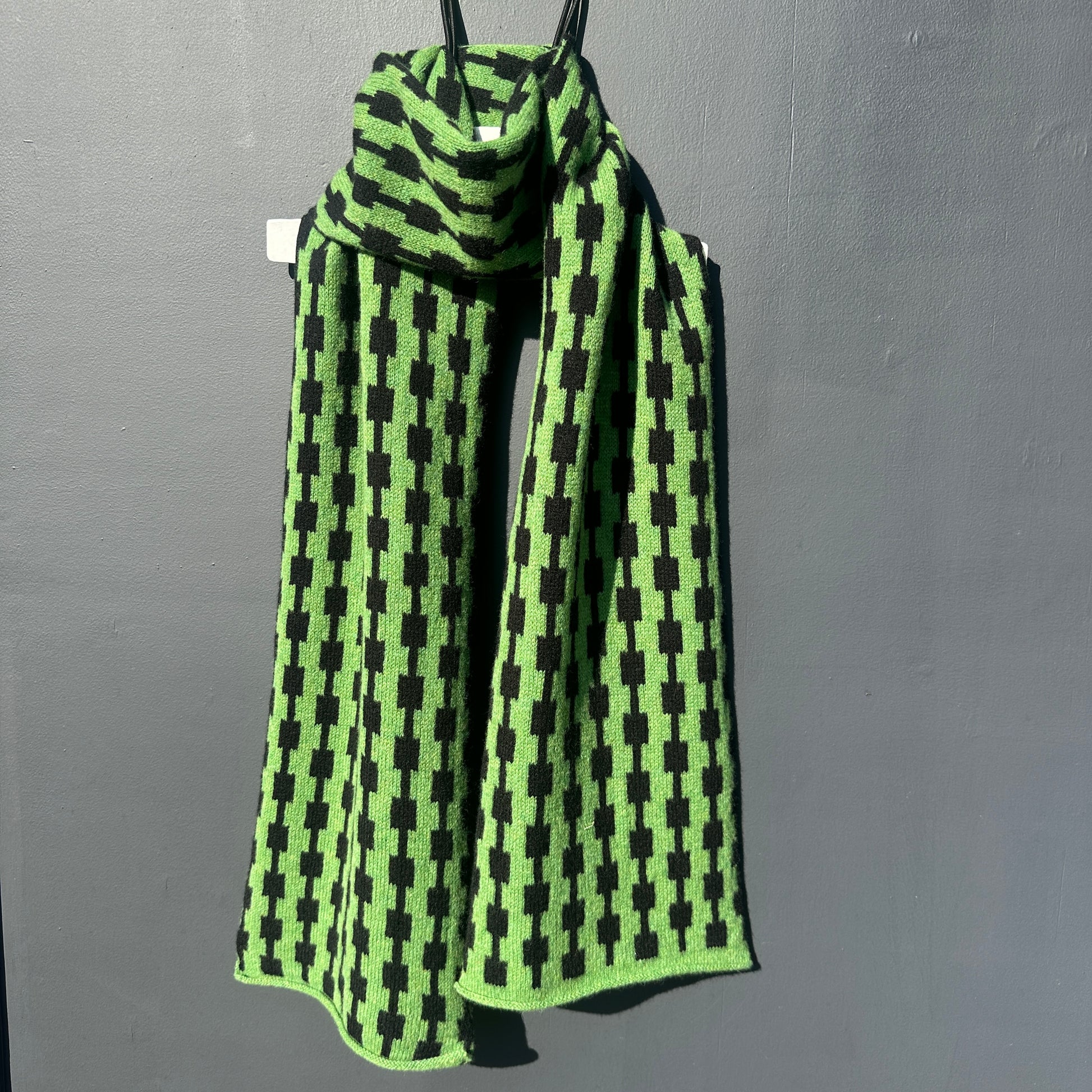 Watercress green and black handcrafted Merino Lambswool geometric patterned Pulse scarf on a white hanger against a grey background.  Different drapping. Pom Shanty Knitwear. 