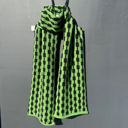 Watercress green and black handcrafted Merino Lambswool geometric patterned Pulse scarf on a white hanger against a grey background.  Different drapping. Pom Shanty Knitwear. 