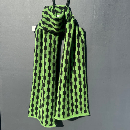 Watercress green and black handcrafted Merino Lambswool geometric patterned Pulse scarf on a white hanger against a grey background.  Different drapping. Pom Shanty Knitwear. 