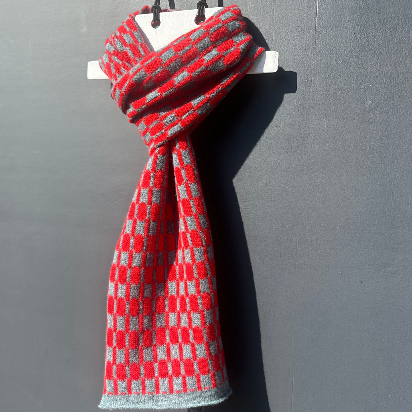 Red and Caspian Blue handcrafted Merino Lambswool geometric patterned Morse scarf on a white hanger against a grey background.  Pom Shanty Knitwear. 