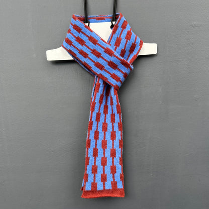 Hanging merino lambswool cornflower blue and sienna brown skinny scarf with a geometric pattern on a grey background. 