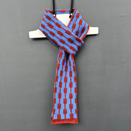 Hanging merino lambswool cornflower blue and sienna brown skinny scarf with a geometric pattern on a grey background. 